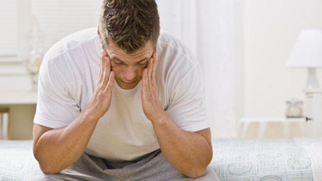 man sitting on edge of bed holding head with both hands due to lack of sleep