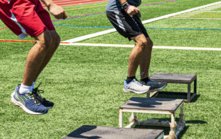 Two male high school athletes doing box jumps on a green turf field during strength and agility practice.