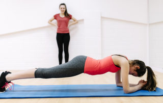 A teenaged girl adopts the 'plank' position in an exercise studio. Her training partner is visible in the back ground.