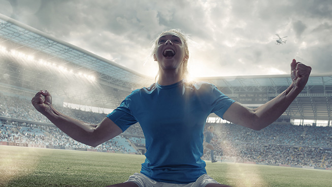 A close up cross processed image of a professional woman soccer player kneeling on the grass with her arms out, fists clenched and head back, shouting in happiness and celebration at winning. The player is in a generic floodlit stadium full of spectators under a cloudy evening sky.