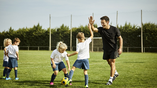 Smiling male soccer coach high-fiving with boy. Children are practicing on field. They are in sports uniforms.