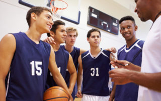 Male High School Basketball Team Having Team Talk With Coach In Gymnasium
