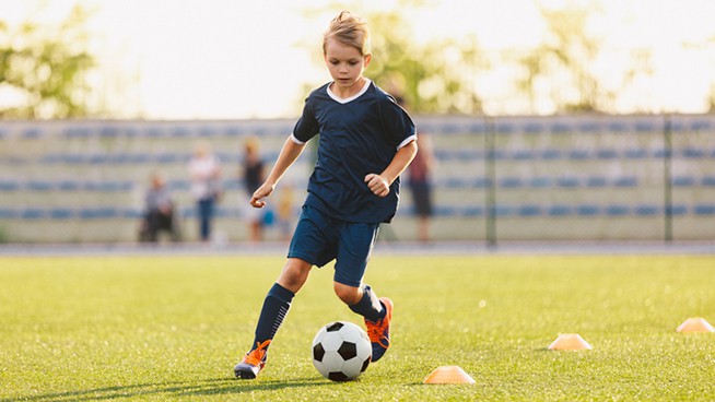 Young boy in blue soccer jersey uniform running after ball on training pitch. Kid improving dribbling skills on practice session