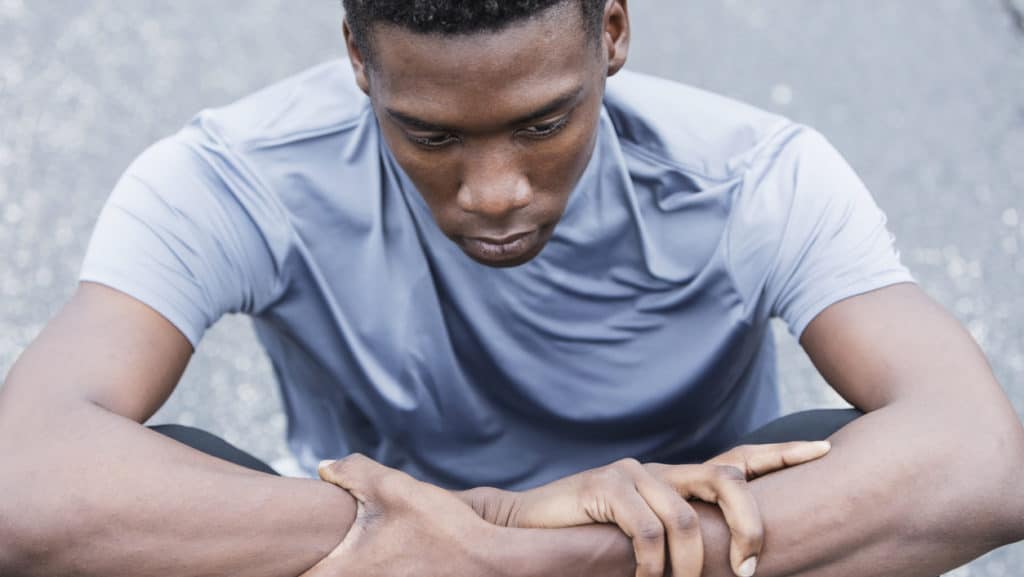 Close-up of a serious young African American man sitting on the pavement. He is an athlete wearing sports clothing, in deep thought, looking down with a serious, sad expression.